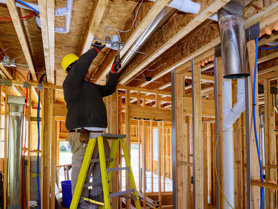 An electrician on a ladder running wire through the framing of a new house